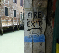 ein lustiger, handgemalter Hinweis zum Feuerausgang in einem Buchladen in Venedig. Ein Pfeil zeigt den Weg in den Kanal, in dem ein Strickmännchen schwimmt